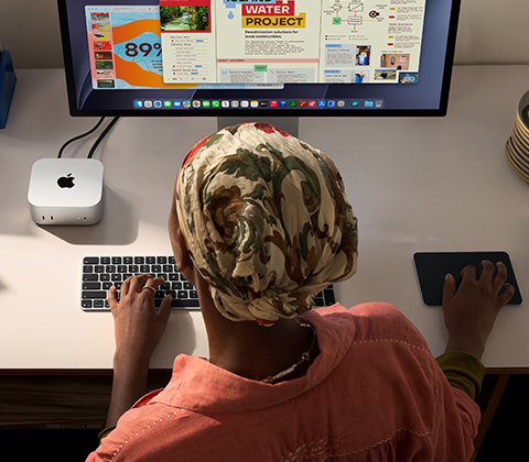 บุคคลหนึ่งกำลังพิมพ์อยู่ที่โต๊ะที่มี Mac mini ตั้งอยู่และเชื่อมต่อ Studio Display, Magic Keyboard, Magic Trackpad และสิ่งของต่างๆ