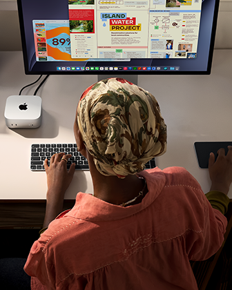 บุคคลหนึ่งกำลังพิมพ์อยู่ที่โต๊ะที่มี Mac mini ตั้งอยู่และเชื่อมต่อ Studio Display, Magic Keyboard, Magic Trackpad และสิ่งของต่างๆ