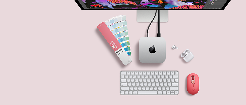 Mac mini, ด้านบน, อะลูมิเนียมสีเงิน, มุมมน, โลโก้ Apple สีดำมันเงาอยู่ตรงกลาง, เชื่อมต่อด้วยสาย, มองเห็น Studio Display ที่ตั้งอยู่ด้านบนบางส่วน, AirPod 4, เปิดฝาอยู่, ตัวเลือกสี Pantone, คีย์บอร์ดและเมาส์ Logitech สีแดง
