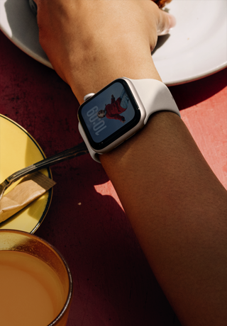 แขนของบุคคลหนึ่งที่เอื้อมไปหยิบอาหารเช้า, Apple Watch SE 3 บนข้อมือ, ตัวเรือนอะลูมิเนียม สีสตาร์ไลท์, จอภาพแบบติดตลอด, ภาพถ่ายของบุคคลบนหน้าปัดนาฬิกา, สายแบบ Sport Band สีสตาร์ไลท์
