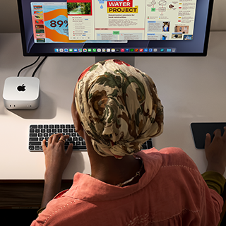 บุคคลหนึ่งกำลังพิมพ์อยู่ที่โต๊ะที่มี Mac mini ตั้งอยู่และเชื่อมต่อ Studio Display, Magic Keyboard, Magic Trackpad และสิ่งของต่างๆ
